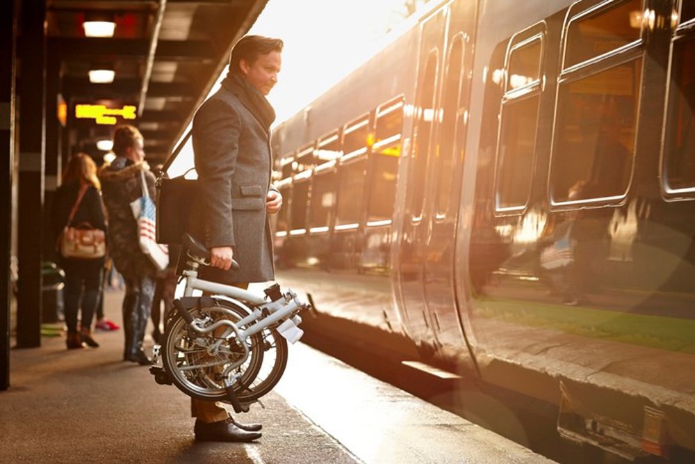 Man standing on station platform beside arriving train with fold up bike in hand