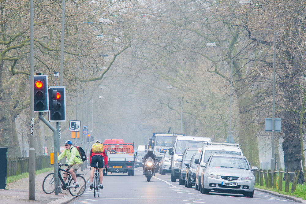 Red traffic lights with traffic building as cyclists cross. The air quality looks slightly hazed