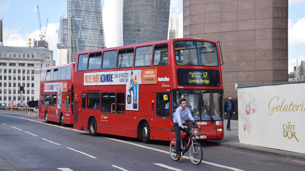 Two red London busses pulled up to road side with man on Boris bike cycling past